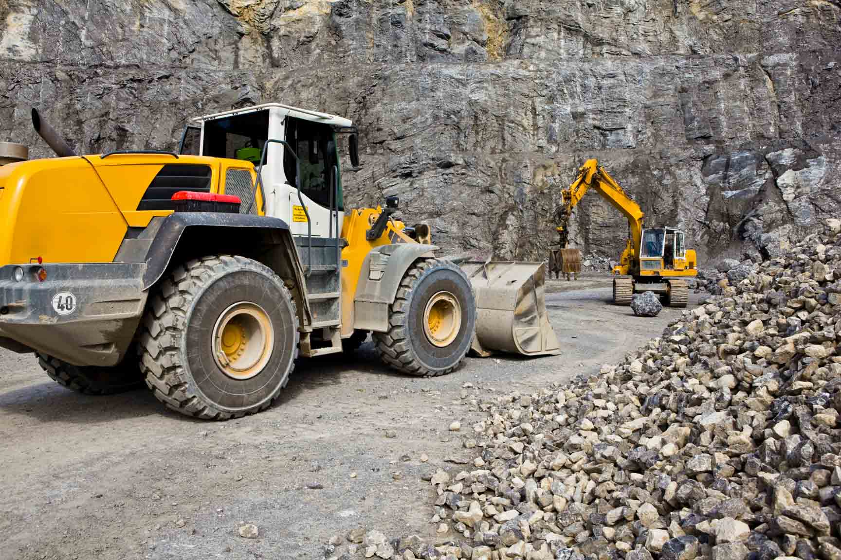 Radlader und Bagger arbeiten in einem Steinbruch vor einer steilen Felswand und bewegen Gesteinsschutt.