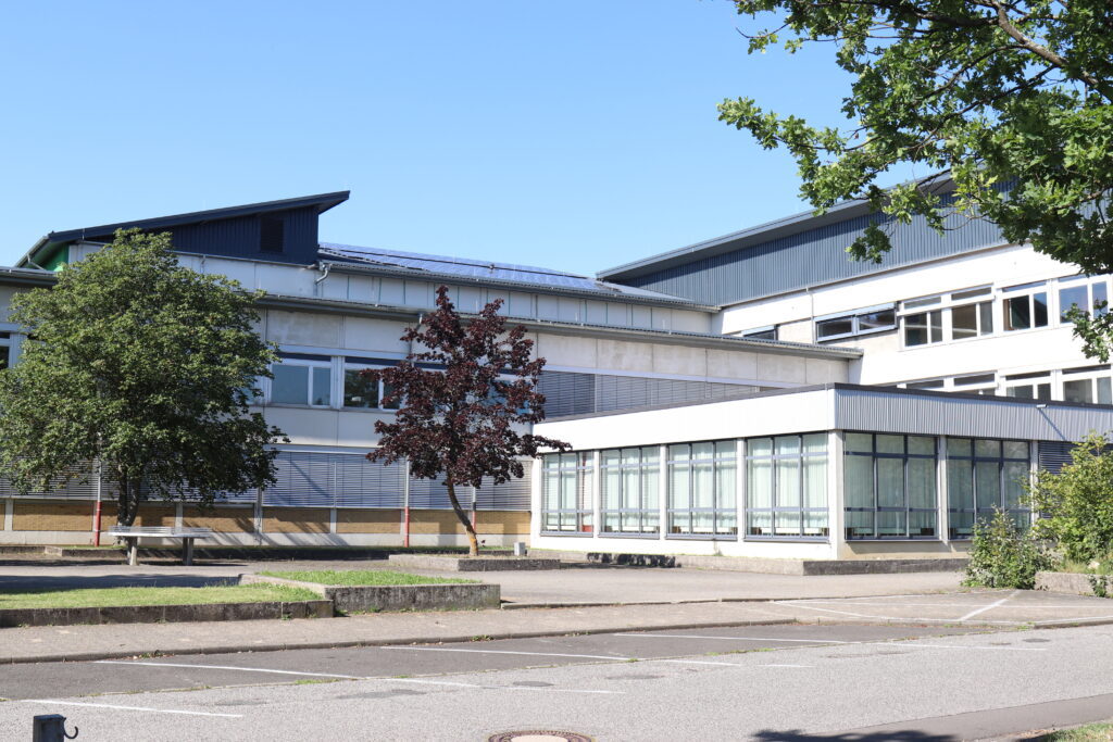 Schulgebäude von außen, davor ein leerer Parkplatz und Bäume.