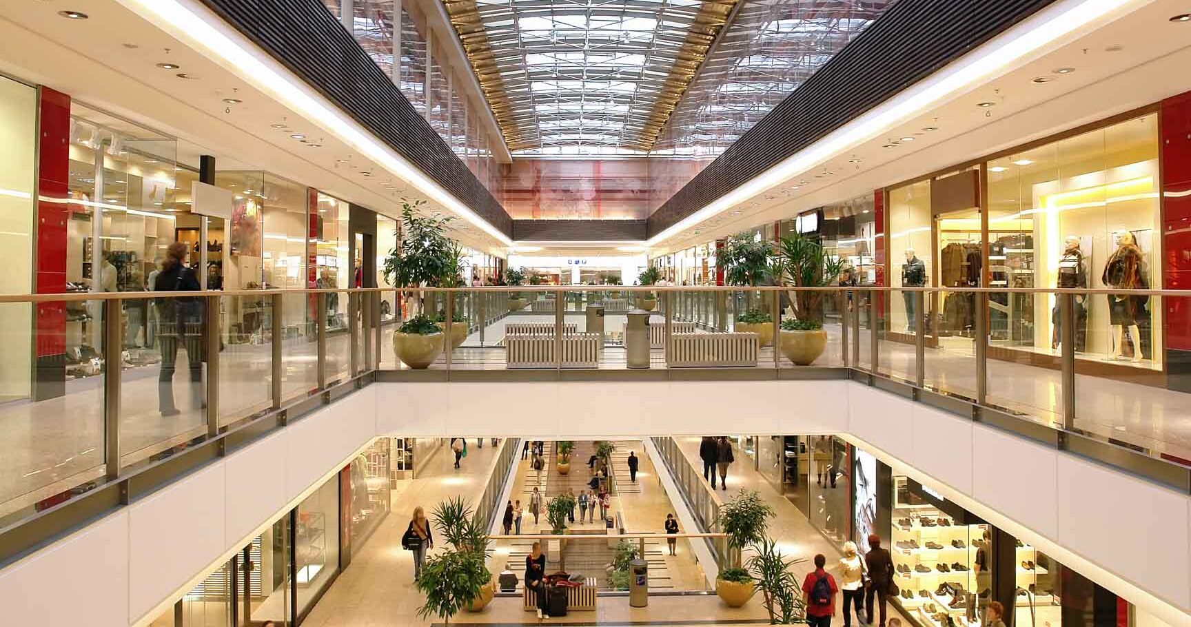 Helles, modernes Einkaufszentrum mit Glasdach, mehreren Etagen, Gesch&auml;ften auf beiden Seiten und Besuchern beim Einkaufen.