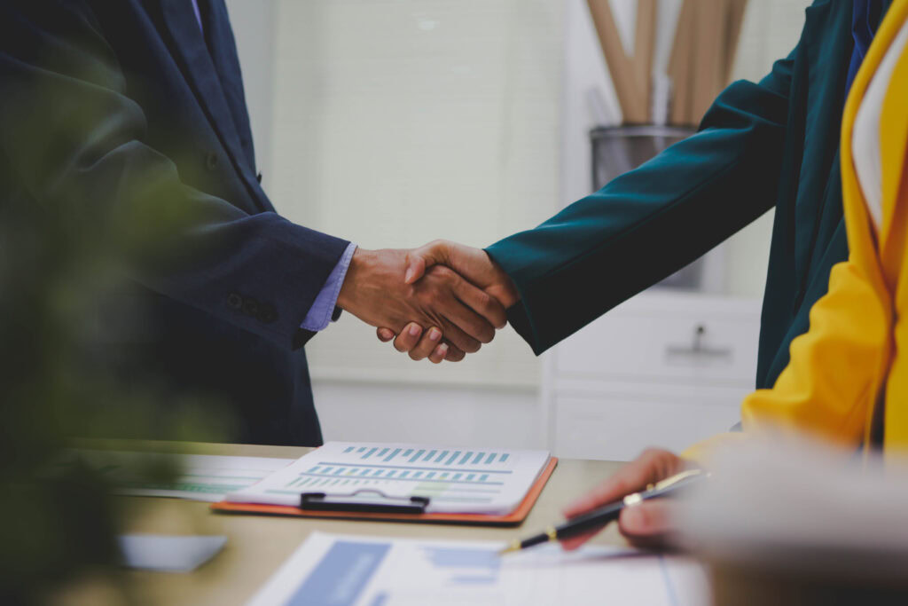 Zwei Personen schütteln sich die Hände, auf dem Tisch nebenbei liegen Unterlagen.