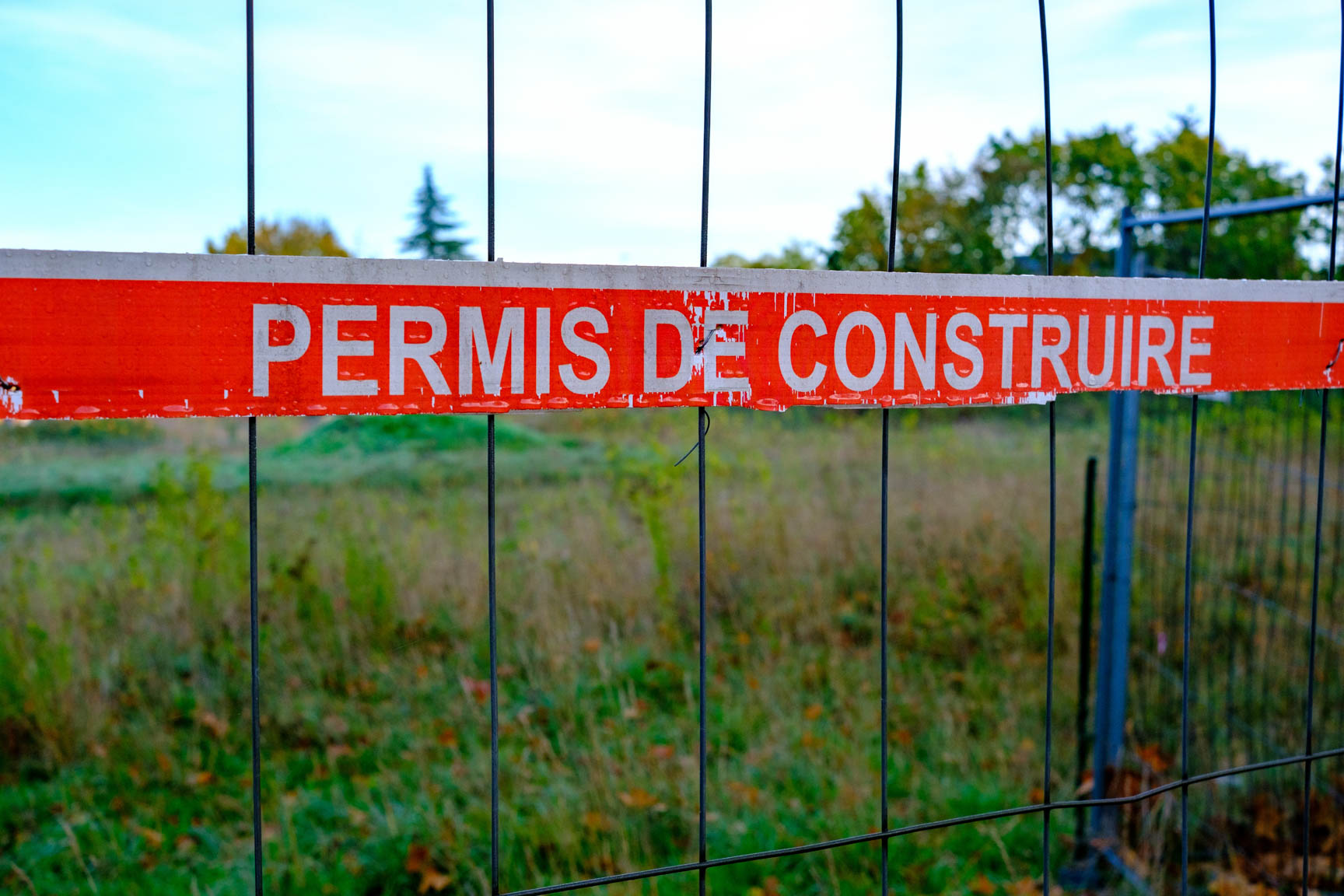 Rotes Absperrband mit der Aufschrift &bdquo;Permis de construire&ldquo; an einem Bauzaun vor einer unbebauten Gr&uuml;nfl&auml;che.