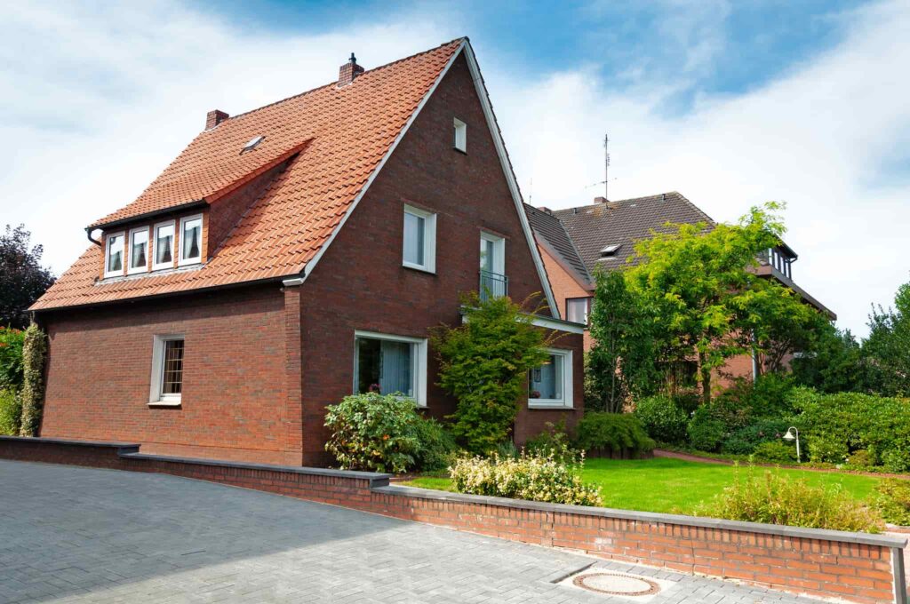 Ein Wohnhaus aus rotem Backstein mit Ziegeldach und grünem Vorgarten, wie es bei Zugewinngemeinschaft und Erbe im Nachlass stehen kann.